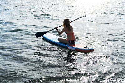 Young woman from behind sitting and paddling on a stand up paddle board on blue sea, sup