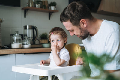 Happy father young man feeds baby girl little daughter in kitchen at home