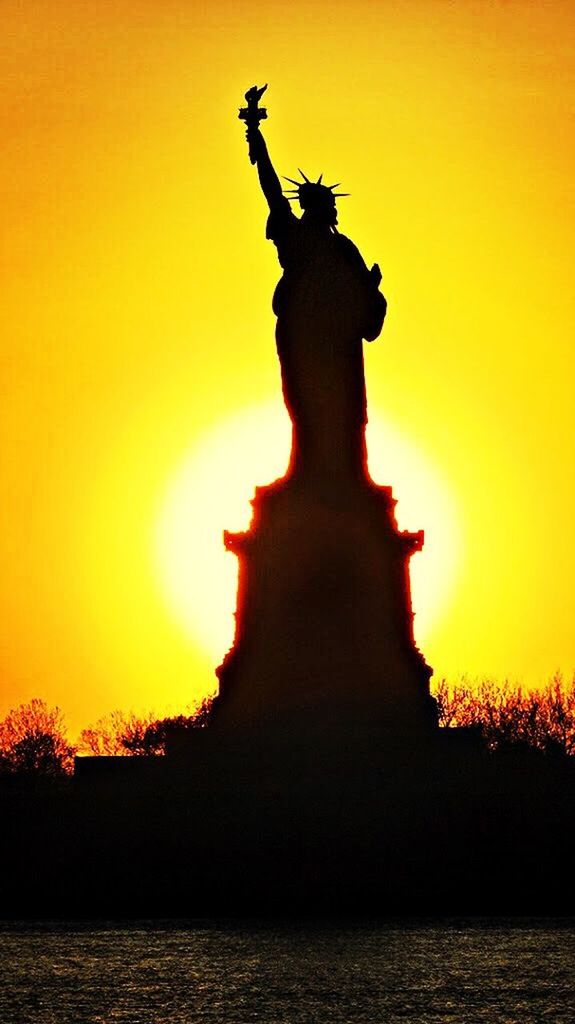 statue, human representation, sculpture, sunset, art, art and craft, creativity, silhouette, orange color, sky, sun, animal representation, low angle view, statue of liberty, outdoors, tourism, water, travel destinations