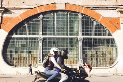 Side view of people riding motorcycle against building on sunny day