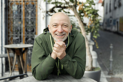 Happy senior man with a beard, clasping his hands joyfully while sitting outdoors in a green jacket