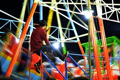 Rear view of man sitting on railing by illuminated amusement park ride at night