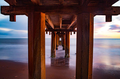 Underneath view of pier on sea against sky during sunset