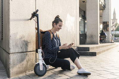 Smiling young woman with electric scooter, earphones and cell phone having a break in the city