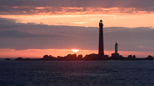 Silhouette lighthouse by sea against cloudy sky during sunset