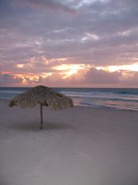 Thatched parasol on sand at beach against cloudy sky during sunset