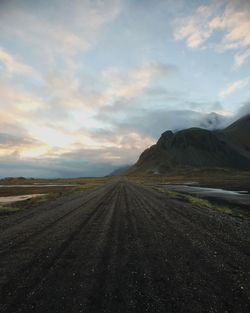 Empty dirt road leading towards mountains against cloudy sky