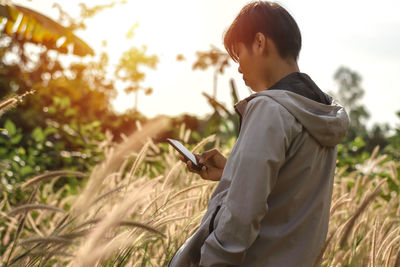 Side view of young woman using mobile phone while standing amidst crops against sky