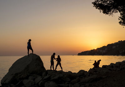 Silhouette people on rocks by sea against sky during sunset