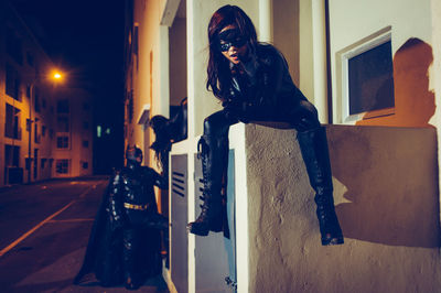 Portrait of young woman wearing superhero costume sitting against building at night