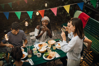 High angle view of young women photographing friends having food during dinner party in backyard