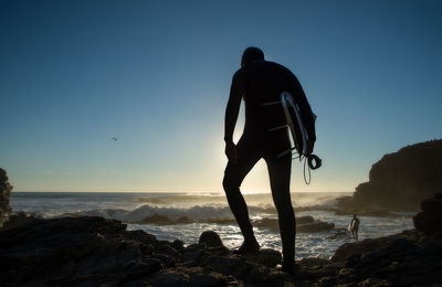 Silhouette man on rock at beach against clear blue sky