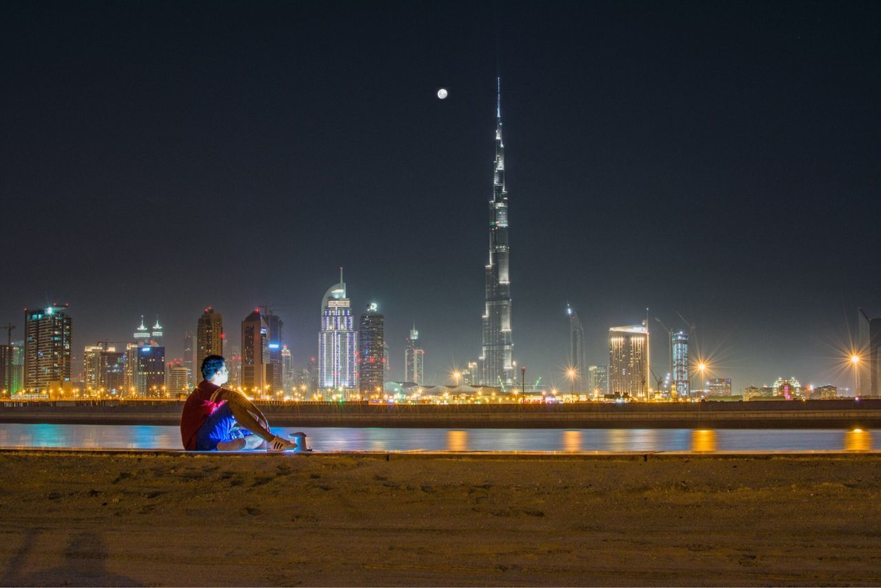 illuminated, night, city, water, architecture, built structure, lifestyles, leisure activity, river, building exterior, men, rear view, full length, standing, cityscape, city life, sky, railing