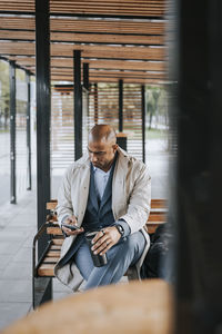 Male business professional using smart phone while sitting on bench at bus stop