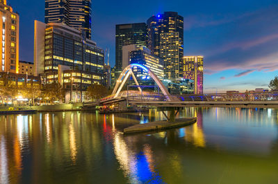 Illuminated bridge over river by buildings against sky at night