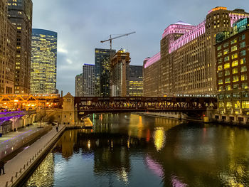 Illuminated bridge over river by buildings against sky at dusk
