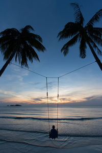 Rear view of person on swing amidst palm trees on beach during sunset