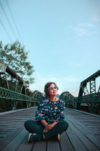 Young woman sitting on footbridge against sky