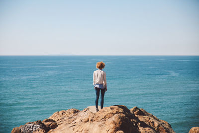 Rear view of woman with curly hair standing on rock at beach against clear sky during sunny day