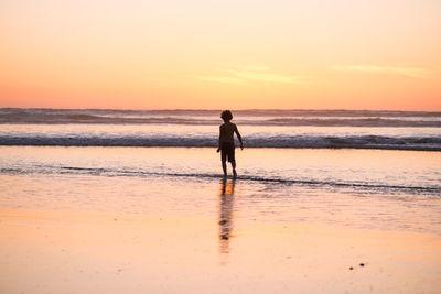 Rear view of shirtless boy walking at beach against sky during sunset