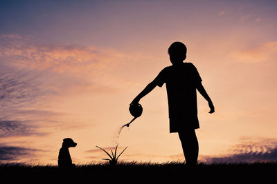 Silhouette of boy watering plant against sky during sunset