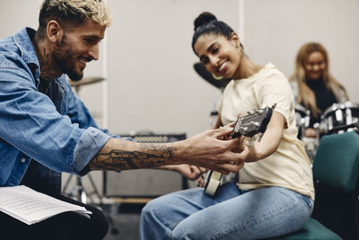 Smiling young man teaching woman guitar while rehearsing in classroom