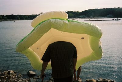 Rear view of man with pool raft standing by lake against sky