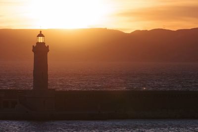 Silhouette lighthouse by sea against sky during sunset