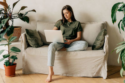 A young attractive brunette girl sitting on a sofa uses a laptop to work remotely online.