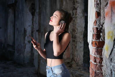 Young woman listening music while standing at abandoned building