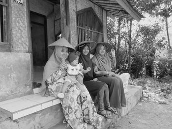 Ladies and a baby in a traditional indonesian bamboo farmer's hat sits in front of an old house