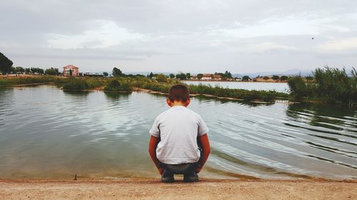 Rear view of boy crouching on riverbank against sky