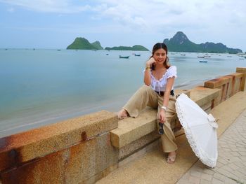 Portrait of smiling young woman sitting by sea against sky