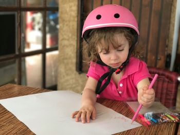 Cute girl wearing pink helmet while drawing on paper at table