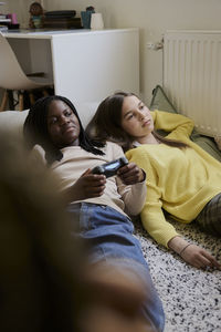 Teenage girl playing video game while lying down with female friend in bedroom