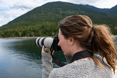 Portrait of woman photographing by lake against mountains