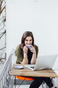 Portrait of smiling young woman with laptop on table having coffee at balcony
