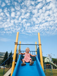 Full length of girl sitting on slide at playground against sky