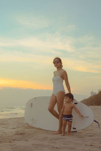 Full length of mother standing with boy with surfing board on beach