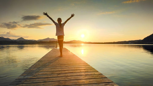 Rear view of woman with arms outstretched standing on pier over lake during sunset