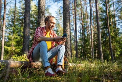 Happy middle aged man browsing in smartphone surfing online sitting on log in park forest.