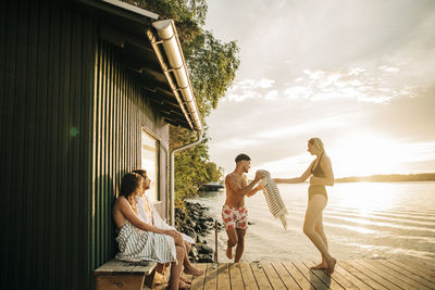 Woman passing towel to male friend while spending vacation at lake during sunset