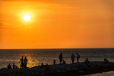 Silhouette people on groyne amidst sea against sky during sunset