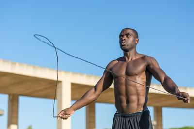 Shirtless man exercising with jump rope against clear sky