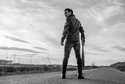 Rear view of mid adult man holding barbed wire wrapped baseball bat while standing on road against sky