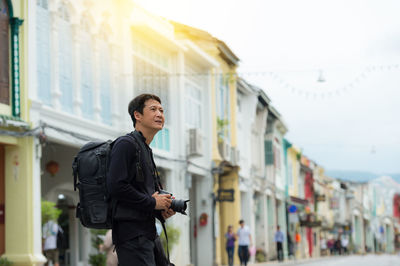 Side view of backpack man with camera standing against buildings in city