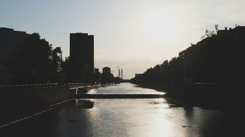 Silhouette buildings by river against sky during sunset