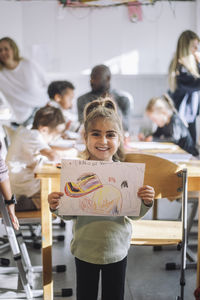 Portrait of smiling girl showing drawing while standing near bench in classroom at kindergarten