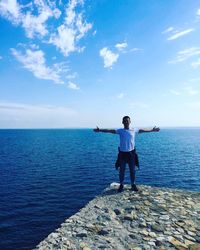 Young man with arms outstretched standing on cliff by sea