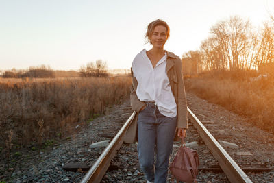 A young woman in a white shirt, beige raincoat and jeans enjoys nature, walking along the railroad 